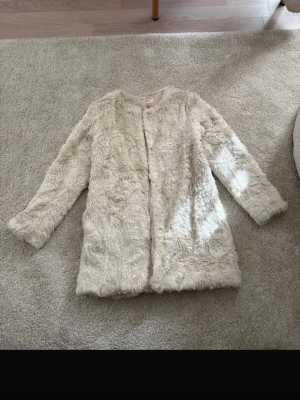 Barn fuskpälsjacka - Säljer en krämvit fuskpälsjacka med rak passform och rund halsringning. Jackan har långa ärmar och stängs framtill. Perfekt för dig som vill ha en mysig och trendig look under kalla dagar.