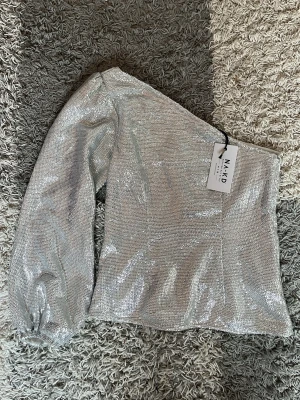 Glittrig oneshoulder topp från NA-KD - Säljer en superglittrig silverfärgad topp från NA-KD med oneshoulder-design och lång ärm. Perfekt statement-plagg med glittrigt material som verkligen sticker ut. Snygg passform och trendig look för dig som vill synas.