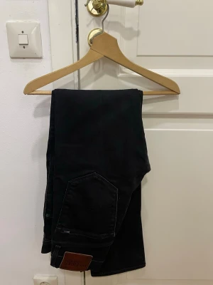 Svarta jeans från Hilfiger Denim - Snygga svarta jeans från Hilfiger Denim med klassisk femficksdesign och läderpatch bak. Jeansen har rak passform och är tillverkade i mjukt bomullsmaterial med lite stretch för extra komfort. Perfekta för en cool och avslappnad stil.