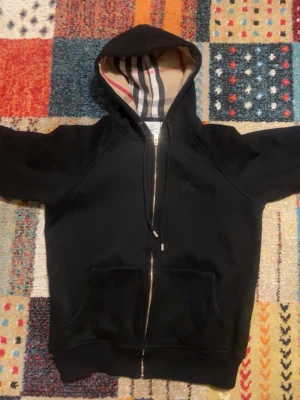 Svart kofta från Burberry -  svart hoodie från Burberry med kedja och klassiskt rutigt foder i huvan. Hoodien har två fickor framtill och en diskret broderad Burberry-logga på bröstet. Perfekt för dig som gillar stilrena plagg med exklusiva detaljer.