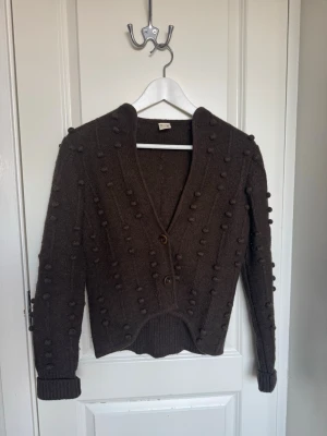 Brun kofta från Rutzou - Mysig brun kofta från Rutzou med dekorativa bollar över hela plagget. Koftan har v-ringning, två knappar framtill. 