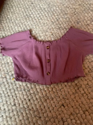 Ljuslila croppad offshoulder topp - Säljer en ljuslila croppad topp med offshoulder-design och ribbad struktur. Toppen har korta ärmar och knappar framtill som snygg detalj. Perfekt för dig som gillar trendiga och bekväma plagg med lite edge.