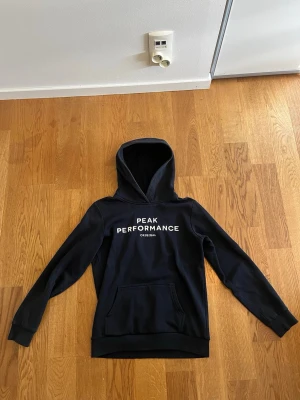 Mörkblå hoodie från Peak Performance - Snygg svart hoodie från Peak Performance med vit logga på bröstet. Klassisk modell med huva och stor magficka framtill. Tillverkad i mjukt material. Storlek 160. 