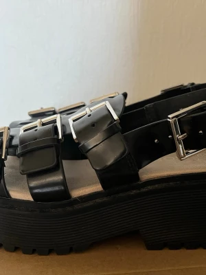 Chunky svarta sandaler med spännen - Säljer ett par chunky svarta sandaler med breda remmar och stora silvriga spännen. Sandalerna har en rejäl platåsula och är tillverkade i konstläder. Perfekta för dig som gillar en edgy och trendig stil.