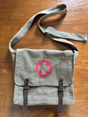 Militärgrön axelväska med rött kors från Rothco - Militärgrön Rothco axelväska med justerbar axelrem och två bruna spännen i läder. På framsidan finns ett tryckt rött kors i cirkel. Vintage army-stil och knappt använd!