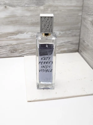 Katy Perry's Indi Visible parfym 100ml 85% kvar - Katy Perry's Indi Visible parfym i en rektangulär glasflaska med silverfärgat lock och svart text på framsidan. Flaskan har en modern och stilren design med transparent glas som visar den ljuslila vätskan inuti.