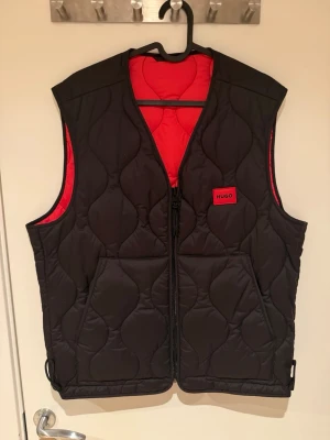 Svart och röd dunväst från Hugo - Stilren svart dunväst från Hugo med röd insida och quiltat mönster. Västen har två stora fickor framtill, dragkedja och en tydlig Hugo-logga på bröstet. Perfekt för lager-på-lager och ger en cool kontrast med den röda fodringen.
