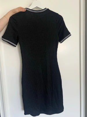 Svart ribbad kortklänning H&M - Svart ribbad kortklänning från H&M med vita ränder på krage och ärmslut. Klänningen har en figurnära passform och korta ärmar, perfekt för en stilren och sportig look.