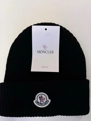 Svart Moncler mössa i ull - Ribbstickad svart mössa från Moncler med uppvikt kant och klassisk Moncler-logga framtill. Tillverkad i mjuk ull som håller dig varm och stilsäker under kyliga dagar. Perfekt accessoar för dig som vill ha både värme och ikonisk design.