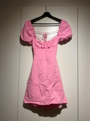 Rosa kort puffärmad klänning - Superfin rosa kortklänning med puffärmar och snörning i ryggen. Klänningen har en söt v-ringning och markerad midja som ger en feminin siluett. Perfekt för dig som vill sticka ut med en färgglad och trendig look.