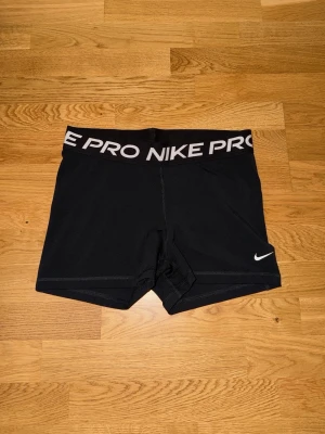 Svarta Nike Pro träningsshorts - Säljer ett par svarta Nike Pro shorts med hög midja och bred resår med Nike Pro-logga. Tight passform och korta ben, perfekta för träning. Tillverkade i stretchigt syntetmaterial för maximal rörelsefrihet.