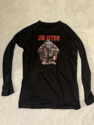 Svart Jiu Jitsu långärmad tröja - Cool svart långärmad tröja med tryck av en apa i Jiu Jitsu-dräkt och texten 'JIU JITSU' i rött. Materialet är troligtvis polyester eller liknande funktionsmaterial, perfekt för träning eller kampsport. Unik design med detaljerade illustrationer och sportig känsla.