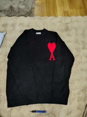 Svart stickad tröja från Ami paris - Snygg svart stickad tröja från Ami med rund halsringning och ribbade muddar. På bröstet finns ett stort rött hjärta med bokstaven A, vilket ger en unik och trendig look. Perfekt för dig som vill ha något stilrent men ändå sticker ut.