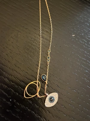 Guldhalsband från Swarovski - Äkta Swarovski halsband med ”onda ögat”. Oanvänt och asken medföljer. Unikt halsband i guldton med flera blå stenar och ett ögonformat hänge dekorerat med små gnistrande stenar. Kedjan har även två cirkelformade detaljeri och små blå infattade stenar längs länken. 