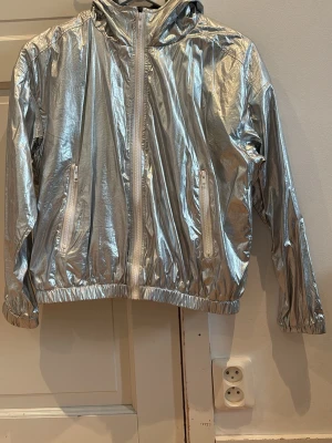 Metallic silver vindjacka med huva - Snygg vindjacka i metallic silver med dragkedja framtill och två fickor med dragkedja. Jackan har elastiska muddar vid ärmslut och nederkant samt en huva. Materialet är glansigt och ger en futuristisk vibe. Perfekt för dig som vill sticka ut.