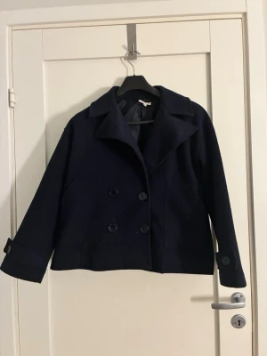 Mörkblå kappa - Stilren mörkblå kappa med dubbelknäppning och bred krage. Jackan har långa ärmar med knappar vid ärmslut och en rak, något croppad passform. Perfekt för dig som gillar klassisk och enkel stil.