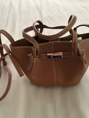  Ny Brun handväska med axelrem - Snygg brun handväska i konstläder med justerbar axelrem och dubbla handtag. Väskan har vita kontrastsömmar och ett dekorativt metallspänne framtill. Perfekt för dig som gillar stilrena accessoarer med en modern touch., Snabb leverans 