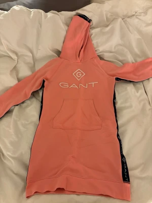 Rosa hoodieklänning från GANT - Snygg rosa hoodieklänning från GANT med vit logga framtill och svarta detaljer längs sidorna. Har huva och en stor magficka. Perfekt för en avslappnad och trendig look. Långa ärmar och mjukt material.