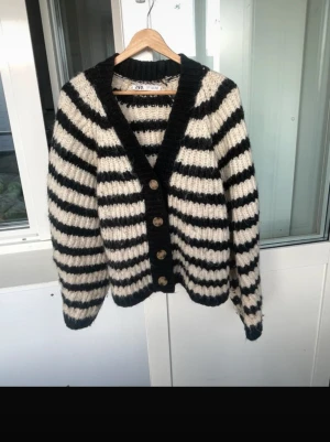 Randig kofta från Zara - Säljer en chunky stickad kofta från Zara med breda svarta och vita ränder. Koftan har djup V-ringning, stora knappar framtill och ribbstickade muddar. Perfekt för lager-på-lager och ger en cool, avslappnad vibe.