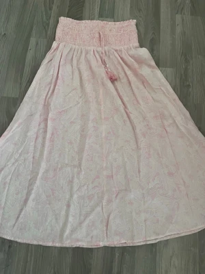 Ljusrosa långkjol med knytband - Söt ljusrosa långkjol med diskret paisleymönster och bred smock i midjan. Kjolen har dekorativa knytband med pärlor och tofsar framtill. Perfekt för sommarens alla tillfällen och ger en bohemisk känsla.