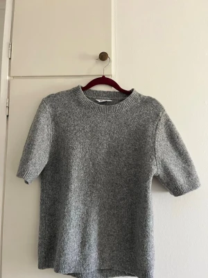 Grå stickad t-shirt i ullblandning - En enkel och stilren grå stickad t-shirt med korta ärmar och rund hals. Tröjan har ribbade muddar vid ärmslut och nederkant. Perfekt för lager på lager och passar till det mesta. Materialet känns mjukt och har en lätt melerad look.