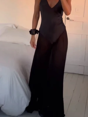 Svart silke långklänning med v-ringning - Elegant svart långklänning med djup v-ringning och ärmlös design. Klänningen har en figurnära överdel och ett flödande, lätt genomskinligt kjolparti som ger en snygg siluett. Perfekt för dig som vill ha en stilren och modern look.