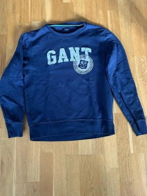 Mörkblå sweatshirt från GANT - Snygg mörkblå sweatshirt från GANT med stort tryck på bröstet där det står GANT och ett emblem under. Tröjan har rund halsringning och ribbade muddar vid ärmslut och nederkant. Perfekt för en avslappnad och stilren look.  Storlek M men sitter som en S/M