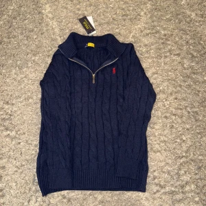 Polo Ralph Lauren Quarter zip  - Mörkblå kabelstickad tröja från Polo Ralph Lauren med halv dragkedja och röd broderad logga på bröstet. Perfekt för lager-på-lager under kyliga dagar. Aldrig använd helt ny. Alla taggar finns kvar aldrig använd. Skickas samma dag!!! Skönt mjukt och tjockt material som passar till vintern.  Storlek S. Quarter zip