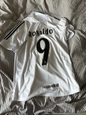 Real Madrid Ronaldo matchtröja - Säljer en vit Real Madrid matchtröja från Adidas med Ronaldo och nummer 9 på ryggen. Tröjan har svarta detaljer, klubbmärke på bröstet och Siemens som sponsor framtill. Perfekt för dig som älskar fotboll och vill ha en klassisk supportertröja.