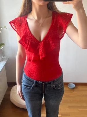 Röd spetsbody med volang från H&M - Supersnygg röd body från H&M i spets med djup v-ringning och volangdetaljer framtill. Ärmlös modell med breda axelpartier och figurnära passform. 