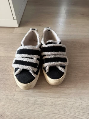Svarta sneakers med teddy från Inuikii - Unika sneakers från Inuikii i svart med fluffiga vita teddy-detaljer och breda snören. Skorna har en chunky beige sula och är fodrade med mjukt teddy-material för extra komfort. Perfekta för dig som vill ha något annorlunda och mysigt på fötterna. Endast använda denna vintern