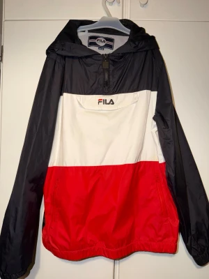  vindjacka från FILA - Snygg vindjacka från FILA i färgblockad design med svart, vitt och rött. Jackan har huva, halv dragkedja framtill, stor ficka på magen och elastisk nederkant. Tillverkad i lätt och tåligt syntetmaterial, perfekt för blåsiga dagar. FILA-logga broderad på bröstet.