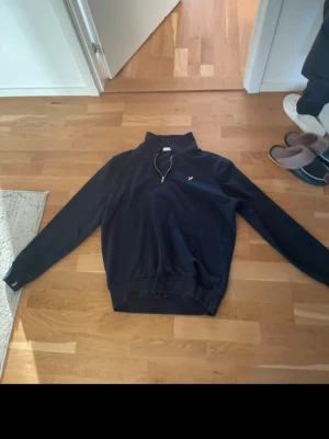 Mörkblå hoodie med halv dragkedja - Snygg mörkblå hoodie från Lyle & Scott med halv dragkedja och gul logga på bröstet. Klassisk passform med ribbade muddar och krage. Perfekt för dig som gillar stilrena och bekväma plagg. Priset kan diskuteras