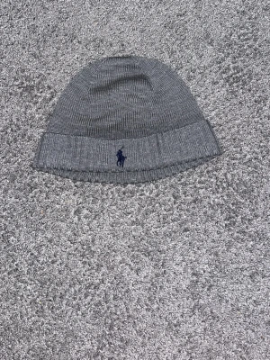 Grå stickad mössa Polo Ralph Lauren - Snygg grå stickad mössa från Polo Ralph Lauren med klassisk ribbad kant och mörkblå broderad logga framtill. Tillverkad i mjukt material som håller dig varm och ger en clean look. Perfekt accessoar för kyliga dagar nu till våren.