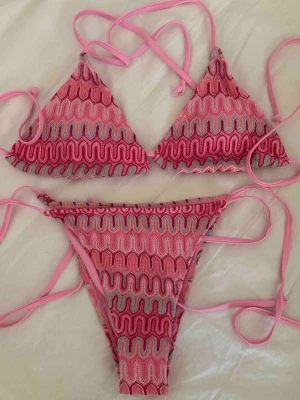 Missoni inspirerad bikini rosa - Trendig trekantsbikini i rosa och ljuslila med vågigt mönster och knytband. Bikinin har en ungdomlig vibe och passar perfekt för strandhäng. Materialet känns mjukt och stretchigt, och toppen har klassisk trekantsform med justerbara band. ENBART TESTAD. Storlek S- passar xs- s. Man får med sån tillhörande kjol som på bilden