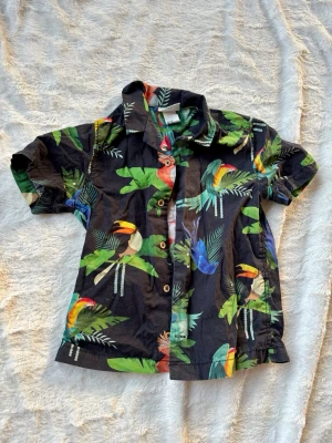 Svart skjorta med tropiskt fågelmönster - Kortärmad svart skjorta med färgglatt tropiskt mönster av papegojor och blad. Skjortan har klassisk krage och knappar framtill. Perfekt för dig som vill sticka ut med en färgstark och somrig vibe.