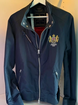 Marinblå jacka från Morris - Snygg marinblå bomberjacka från Morris med broderad logga och emblem på bröstet. En perfekt vårjacka! Storlek S.