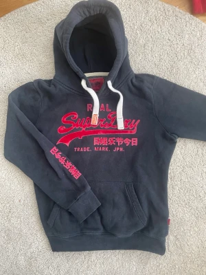 Mörkblå Superdry hoodie med tryck - Mörkblå hoodie från Superdry med röd logga och japansk text på bröstet och ärmen. Klassisk känguruficka framtill, vita dragsnören i huvan och mjukt material. Perfekt för en avslappnad streetwear-look.