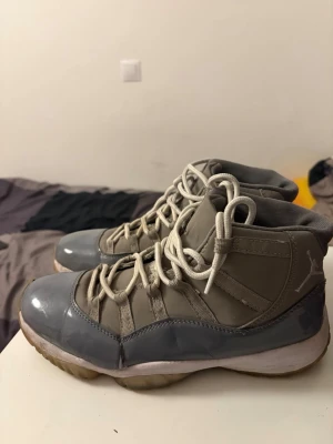 Gråa Air Jordan 11 sneakers - Säljer ett par Air Jordan 11 sneakers i cool grå färg med vita skosnören och genomskinlig sula. Skorna har en blandning av blankt och matt material, med klassisk Jumpman-logga på hälen. Perfekta för dig som gillar retro och streetwear vibes.