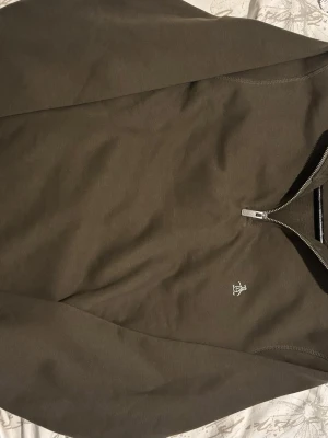 Mörk grön half zip topp från Penguin - Snygg Mörk grön långärmad topp från Penguin med half zip-dragkedja framtill och diskret logga broderad på bröstet. Perfekt för en avslappnad och stilren look. Materialet känns mjukt och bekvämt.