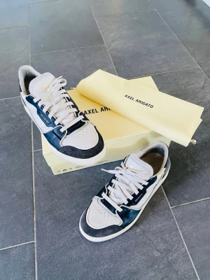 Axel Arigato blåvita sneakers - Snygga sneakers från Axel Arigato i blå och vitt med detaljer i läder och mocka. Klassisk låg modell med vita snören och guldfärgad logga på sidan. Skorna kommer med originalkartong och dustbag för extra premiumkänsla.