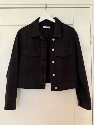 Svart croppad jeansjacka Gina Tricot - Snygg svart croppad jeansjacka från Gina Tricot med klassisk krage, silverfärgade knappar och två bröstfickor med lock. Perfekt till alla outfits för en trendig look.