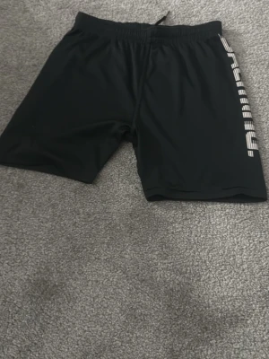 Svarta träningsshorts från Salming - Svarta shorts från Salming med vitt logotryck längs ena benet. De har elastisk midja och är tillverkade i ett lätt och smidigt material som passar perfekt för träning eller sport.