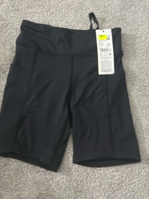 Svarta shorts från Röhnisch - Säljer ett par svarta shorts från Röhnisch med hög midja och tight passform. Shortsen har praktiska sidfickor och är tillverkade i ett stretchigt syntetmaterial som passar perfekt för träning. Stilrena och funktionella för en aktiv livsstil.