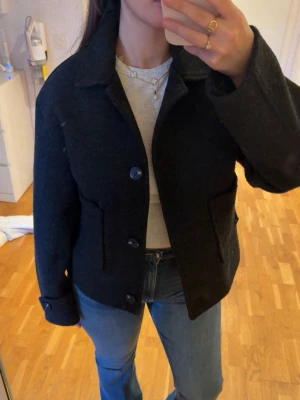 Svart kort jacka med stora fickor - Säljer en svart, kort jacka med klassisk krage och stora fickor framtill. Jackan har 4 knappar och är tillverkad i ett tjockare material som passar bra till kyliga dagar. Perfekt för dig som gillar en stilren och enkel look.