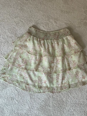 Blommig volangkjol i pastellfärger - Söt kort kjol med blommigt mönster i ljusgrönt, rosa och vitt. Kjolen har flera volanger och en smockad midja för skön passform. Perfekt för dig som gillar romantisk och feminin stil.