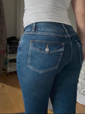 Blå skinny jeans med knappdetalj - Säljer ett par klassiska blå boot cat jeans med hög midja och snygga kontrastsömmar. Jeansen har fem fickor, varav bakfickorna har lock och knappdetalj. Materialet är stretchigt denim som sitter tight och bekvämt. Aldrig använda