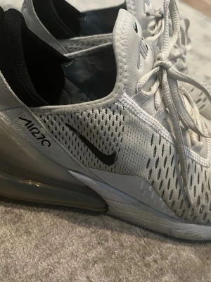 Nike Air Max 270 vita sneakers - Nike Air Max 270 sneakers i vitt med svart Swoosh och AIR270-tryck på sidan. Skorna har en stor synlig Air-enhet i hälen, mesh-ovandel för bra ventilation och klassisk snörning. Insidan är svart och designen är sportig och modern.