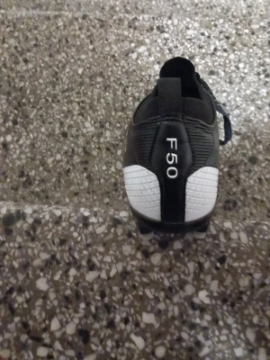 Adidas F50 svart/vit fotbollssko - Säljer en Adidas F50 fotbollssko i svart och vitt med klassisk design och snörning. Skon har en röd innersula och är tillverkad i syntetmaterial för lätt vikt och bra passform. Perfekt för dig som vill ha snabbhet och kontroll på planen.