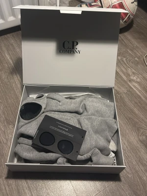 Grå hoodie med goggles från C.P. Company - Säljer en grå hoodie från C.P. Company med dragkedja och deras ikoniska goggles i huvan. Hoodien har en modern passform, ribbade muddar och är tillverkad i mjukt material. Perfekt för dig som vill sticka ut med en unik detalj i streetwear-stil.